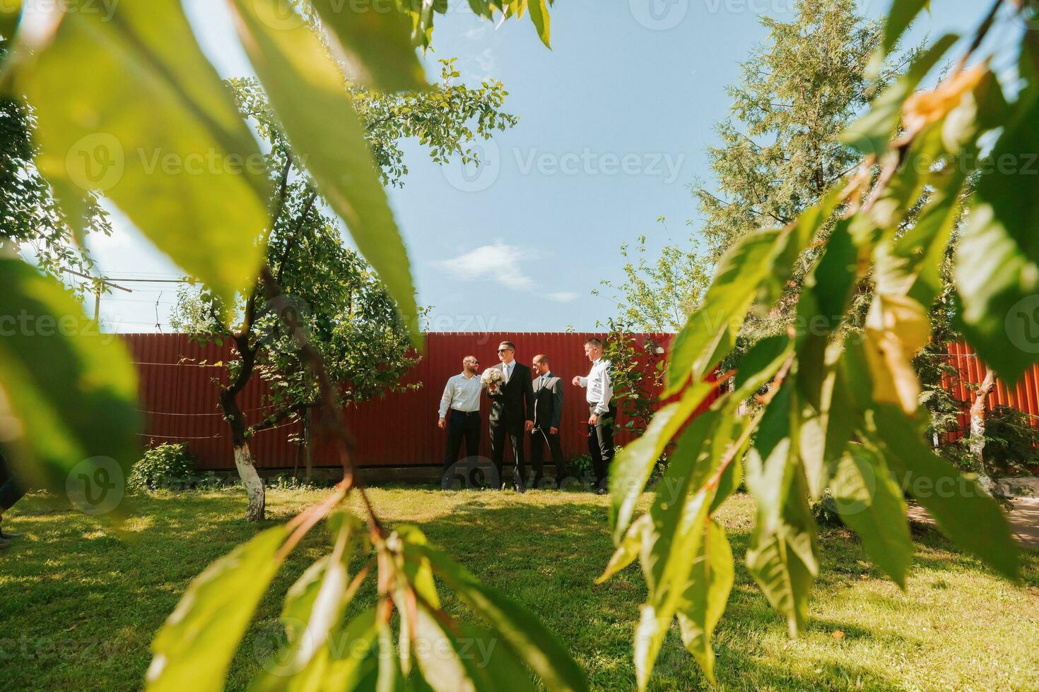 The groom in a black suit and glasses and his stylish friends wearing white shirts and black pants and glasses are standing in the backyard in the garden. The groom is holding a bouquet. photo