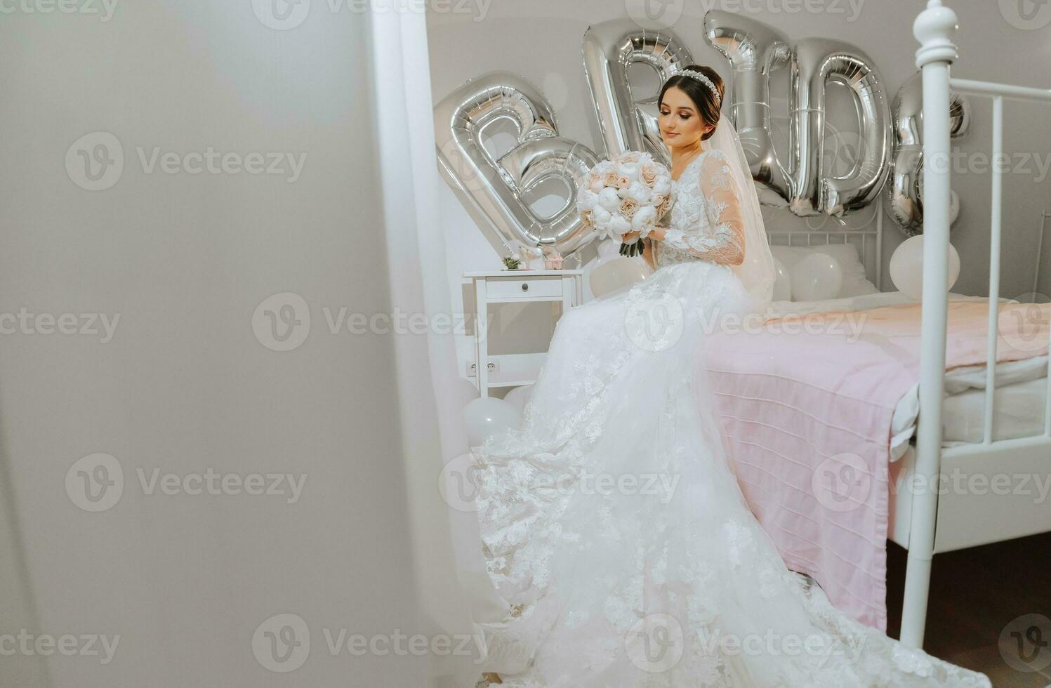 un hermosa joven novia en un Boda vestir con un ramo de flores de peonías en su manos es sentado en un cama en un habitación. final preparativos para el boda. el novia es esperando para el novio. mañana, novia. foto