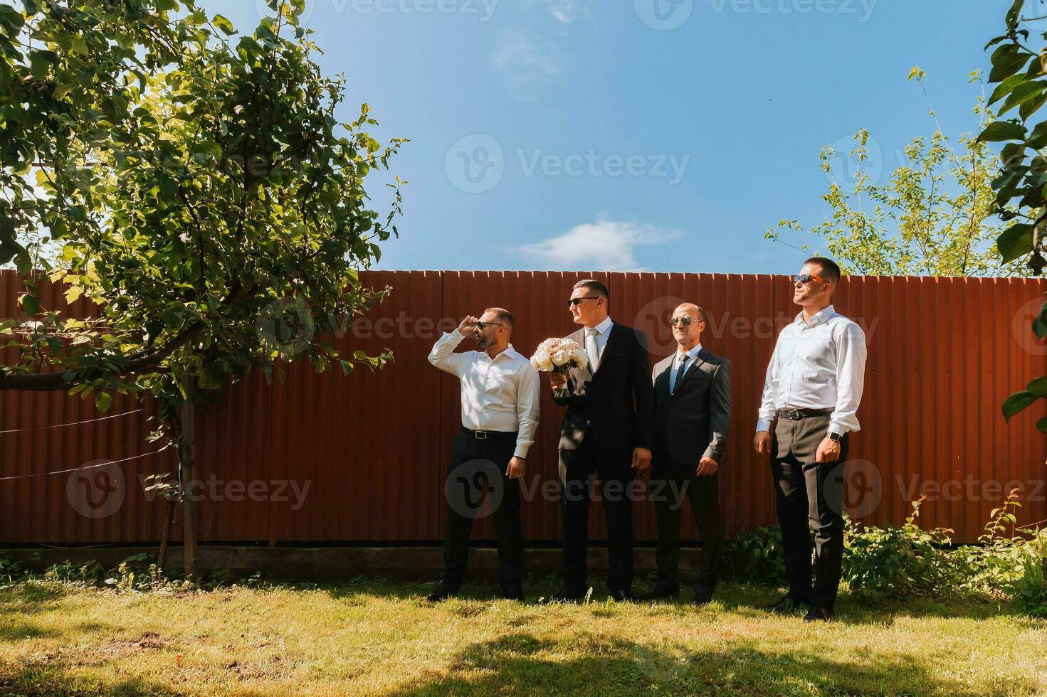 The groom in a black suit and glasses and his stylish friends wearing white shirts and black pants and glasses are standing in the backyard in the garden. The groom is holding a bouquet. photo