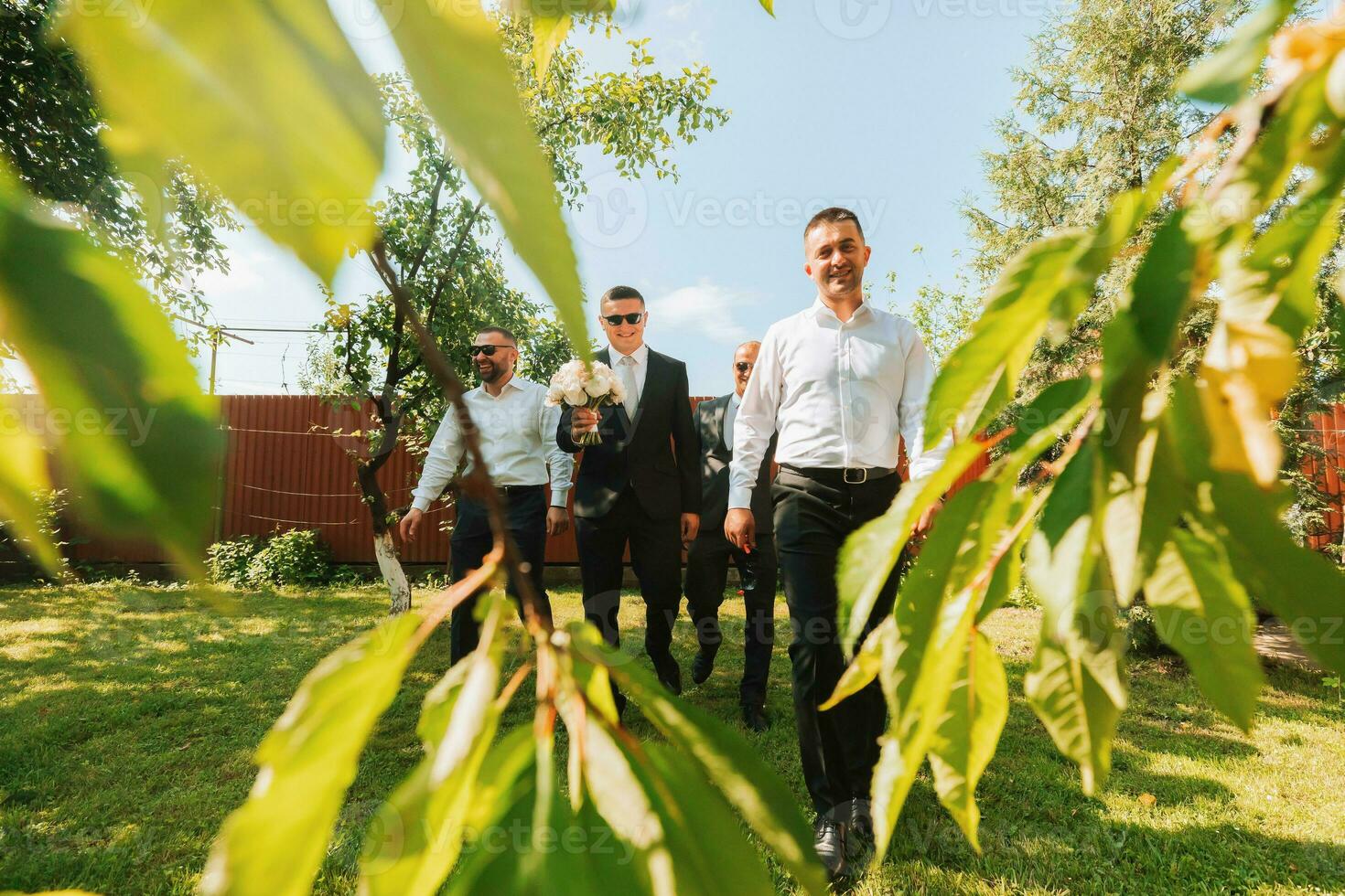 The groom in a black suit and glasses and his stylish friends wearing white shirts and black pants and glasses are standing in the backyard in the garden. The groom is holding a bouquet. photo