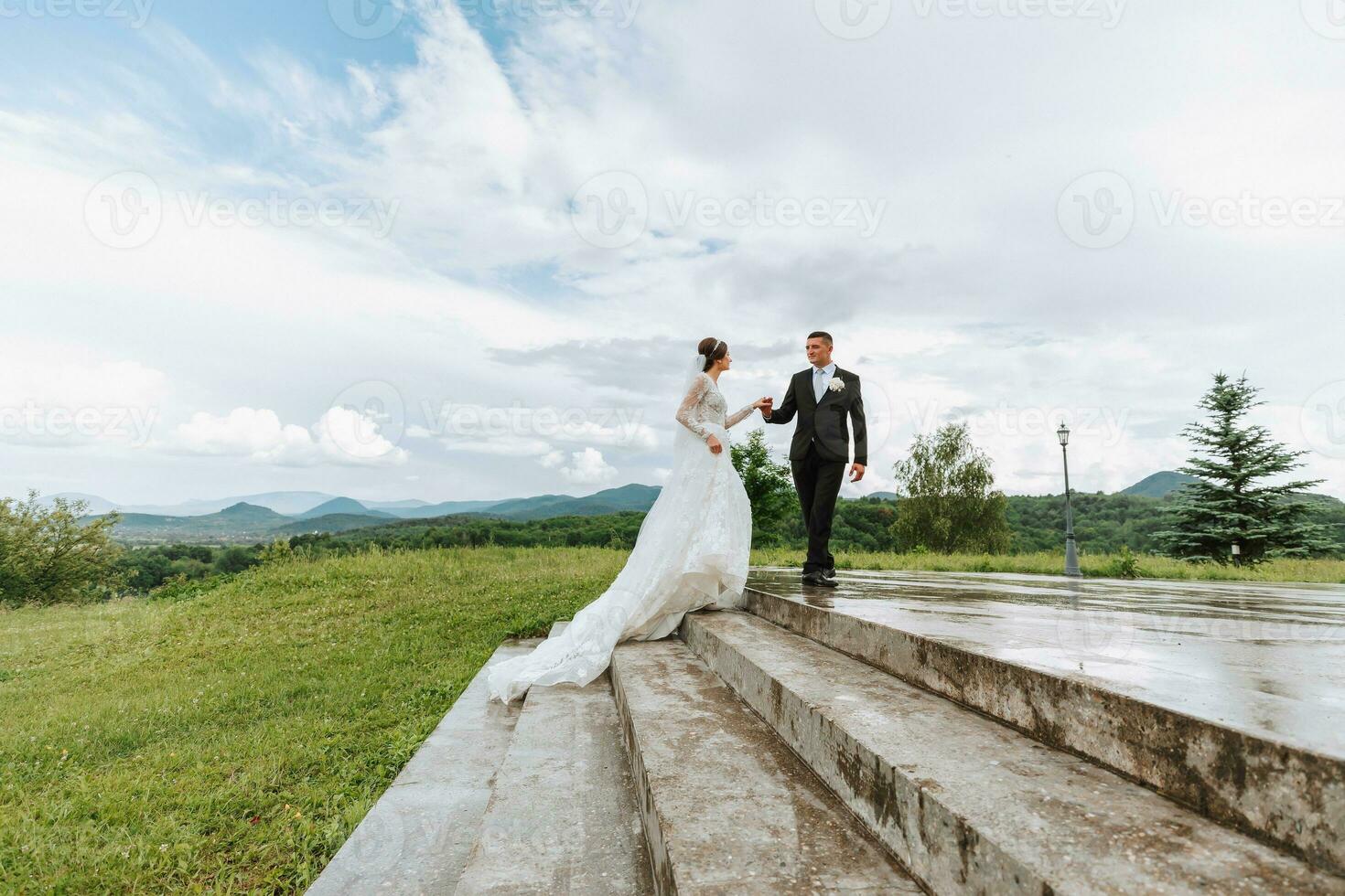 a beautiful bride in a wedding dress with a beautiful hairstyle and a groom in a black suit on the stairs, behind a gorgeous mountain view. Wide-angle photo