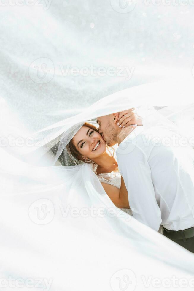 Wedding portrait. The bride and groom covered themselves with a veil, hugged and kissed tenderly. Holiday concept. Transparent veil. Rays of the sun. photo