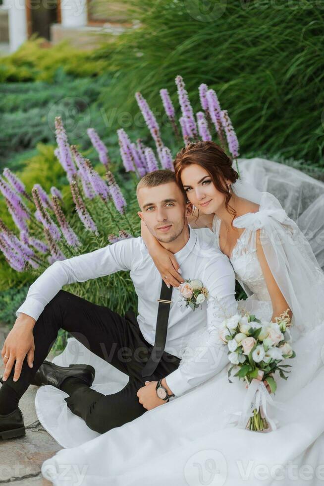 Portrait of the bride and groom in the park. The bride is holding a bouquet and hugging her groom, they are sitting, posing near lavender flowers. Wedding walk in the park. Long train of the dress photo