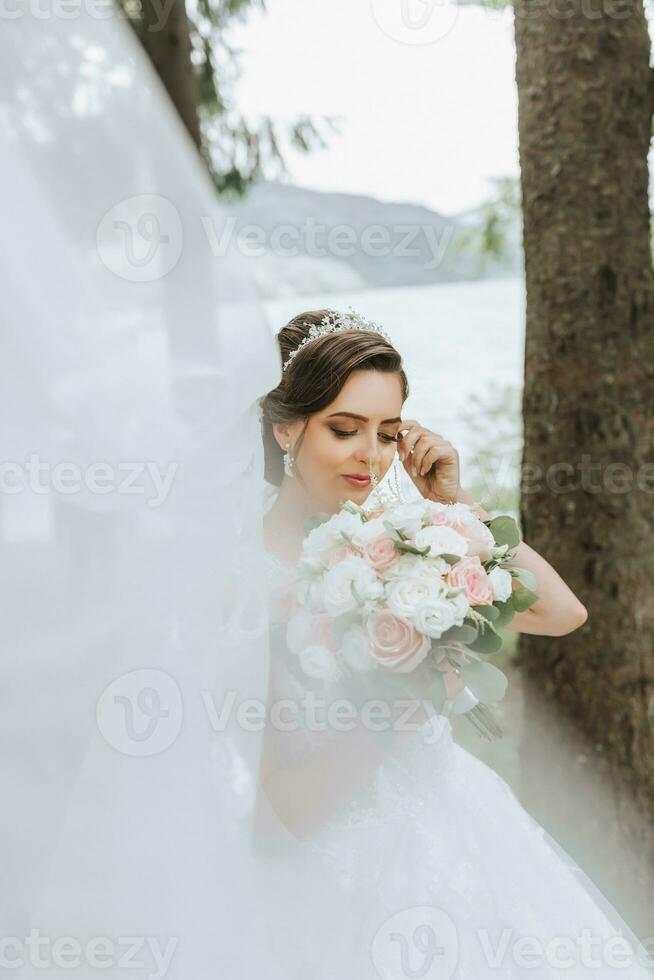 retrato de un novia con un ramo de flores en contra el antecedentes de alto arboles y un lago. un hermosa joven novia es participación un Boda ramo de flores en su manos, el de la novia velo es soplo en el viento. foto