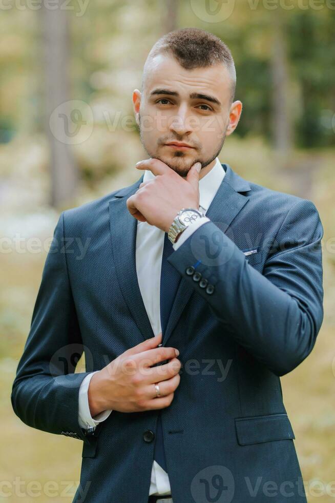 A young man in a classic suit against the background of tall trees in a forest or park. The groom fastens his jacket photo