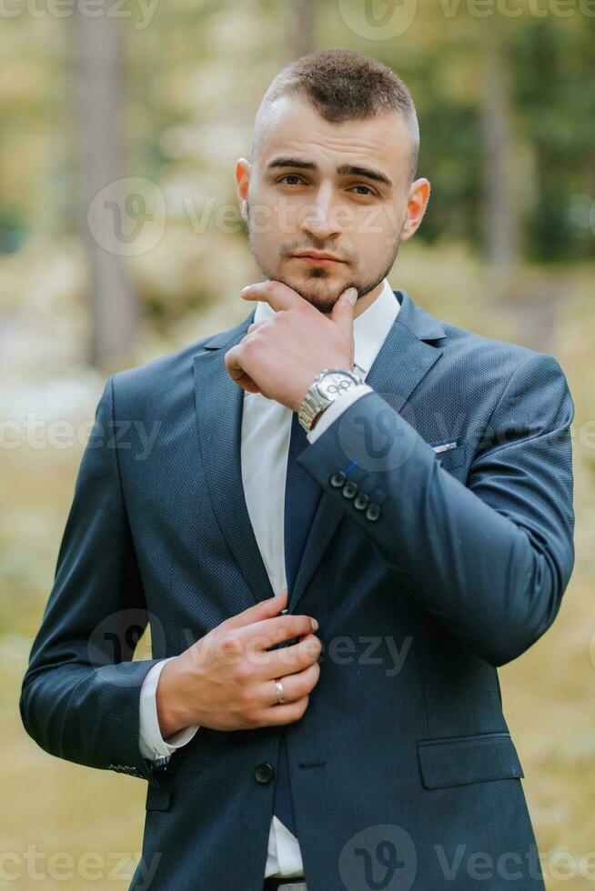 un joven hombre en un clásico traje en contra el antecedentes de alto arboles en un bosque o parque. el novio sujeta su chaqueta foto