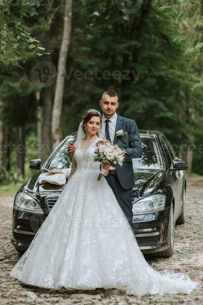 frente ver de un casado novia y novio vistiendo festivo ropa en pie en contra un negro coche en su Boda día foto