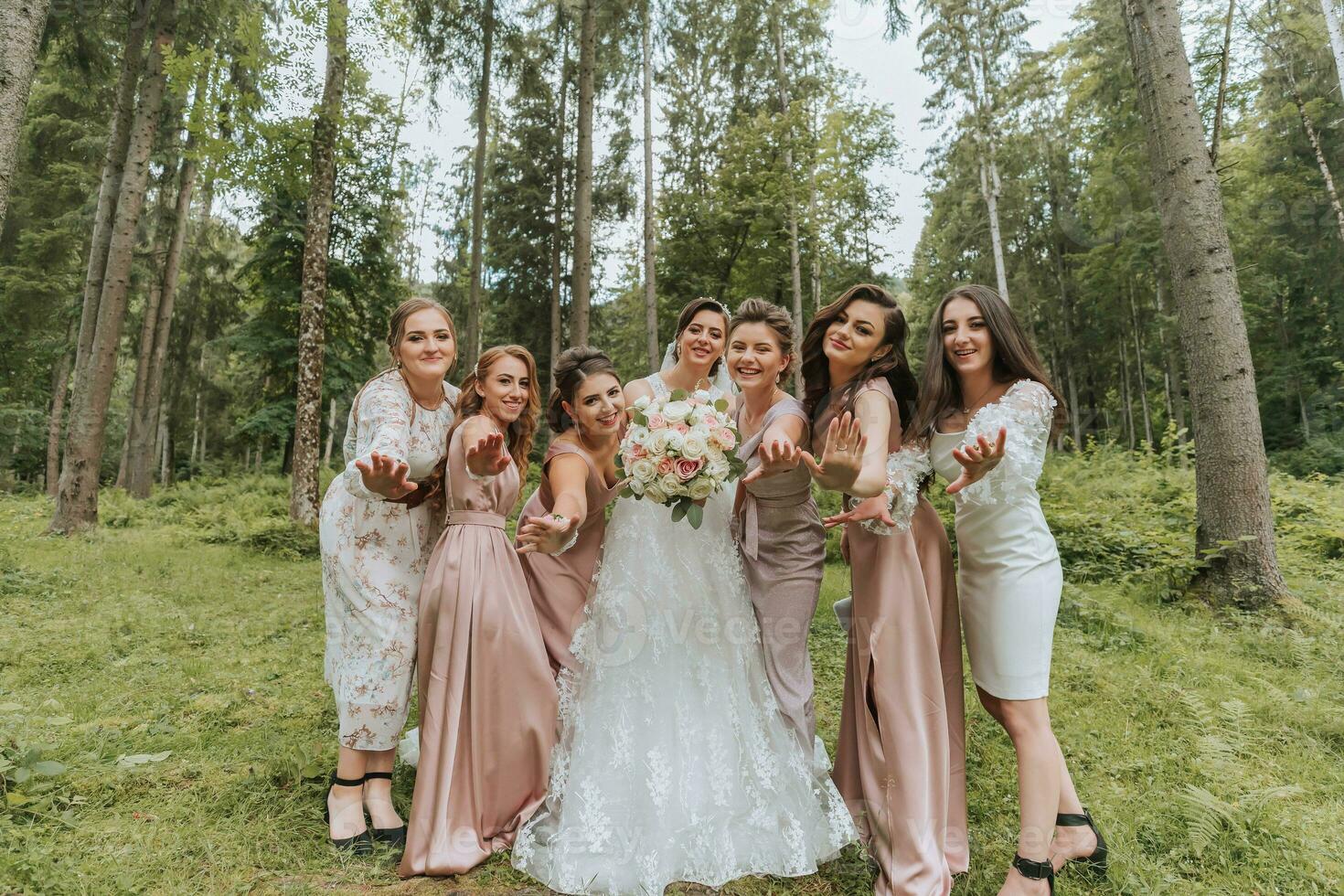 A group of beautiful girls with a bride in identical dresses are smiling, celebrating and having fun together against the background of nature and tall trees. Girls party photo