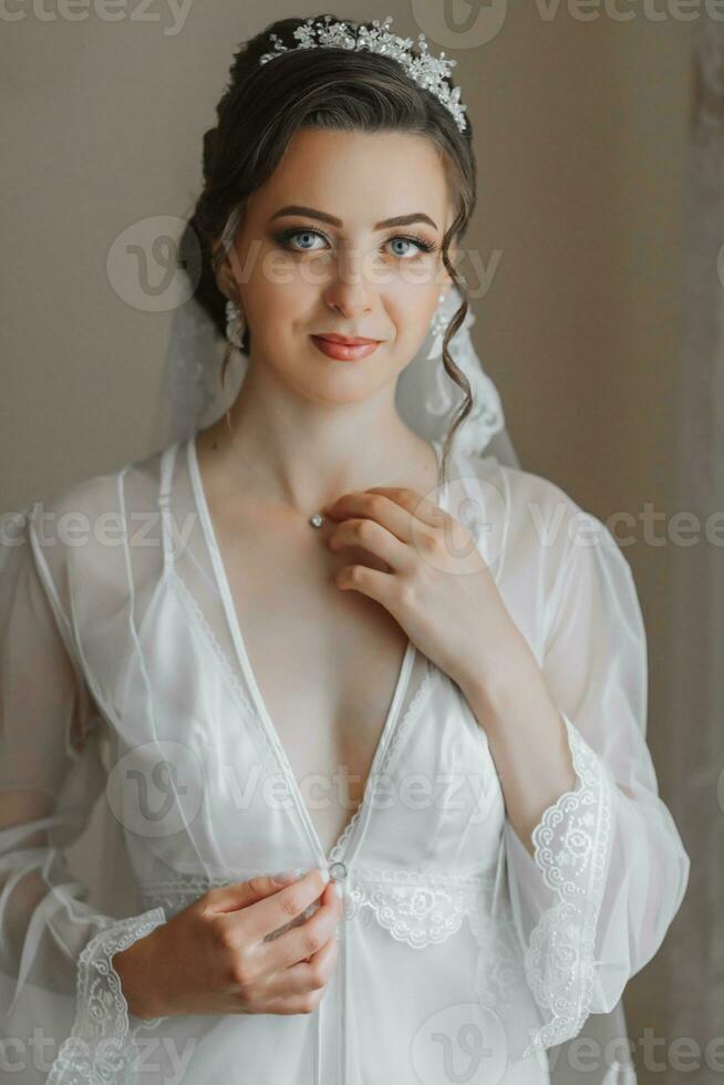 a beautiful bride in a white robe with an open bust touches a diamond pendant on her neck. The bride is preparing for the wedding. bride's fees the wedding day of the newlyweds. photo