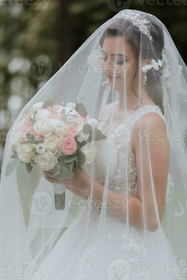 the bride in a wedding dress with a long train and a veil holds a wedding bouquet of roses, under the veil photo