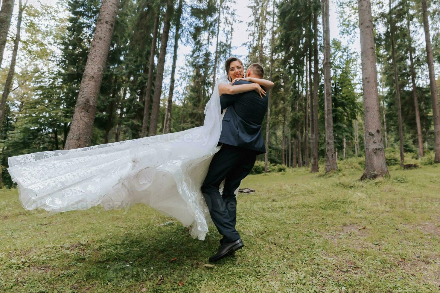 Fashionable groom and cute bride in white dress with long train and crown on head, groom circling with bride in arms, garden, forest outdoors. Wedding photography, portrait of smiling newlyweds. photo