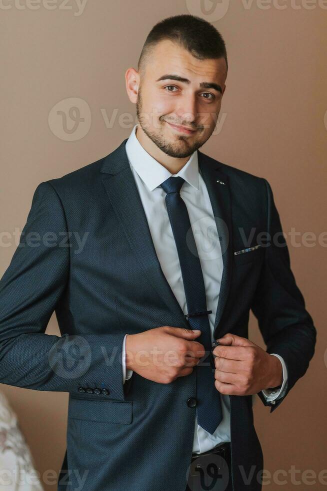 Confident elegant handsome guy standing at home and buttoning his jacket looking at the camera in a nice dark blue suit photo