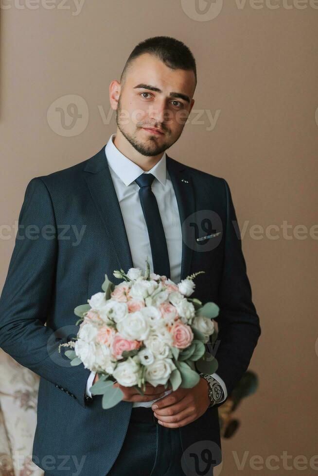 Confident elegant handsome guy in a nice dark blue suit with a bouquet of flowers in his hands. The groom is preparing for the wedding ceremony photo