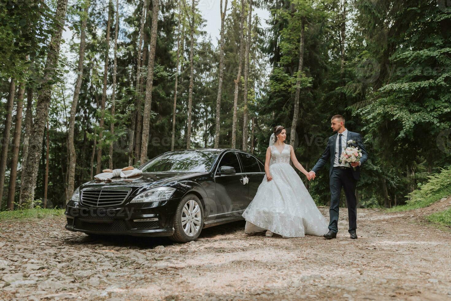 Front view of a married bride and groom wearing festive clothes standing against a black car on their wedding day photo