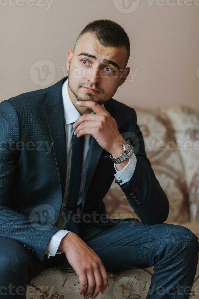 Confident elegant handsome guy at home sitting on the couch in a nice dark blue suit. photo