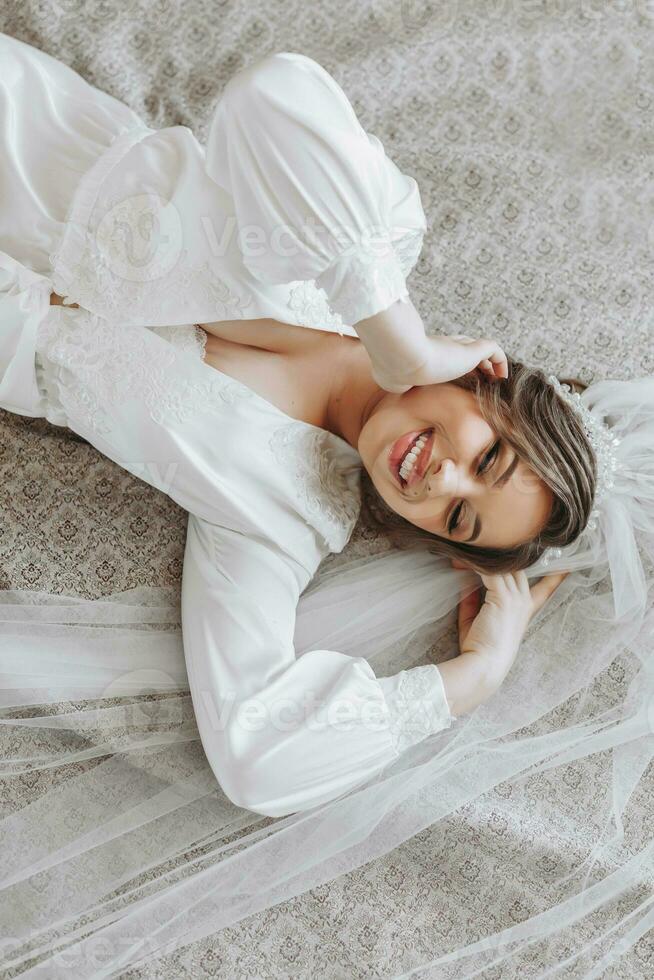 Beautiful young bride in a white peignoir lying on the bed in her bedroom. Final preparations for the wedding. The bride is waiting for the groom. Morning, bride. photo