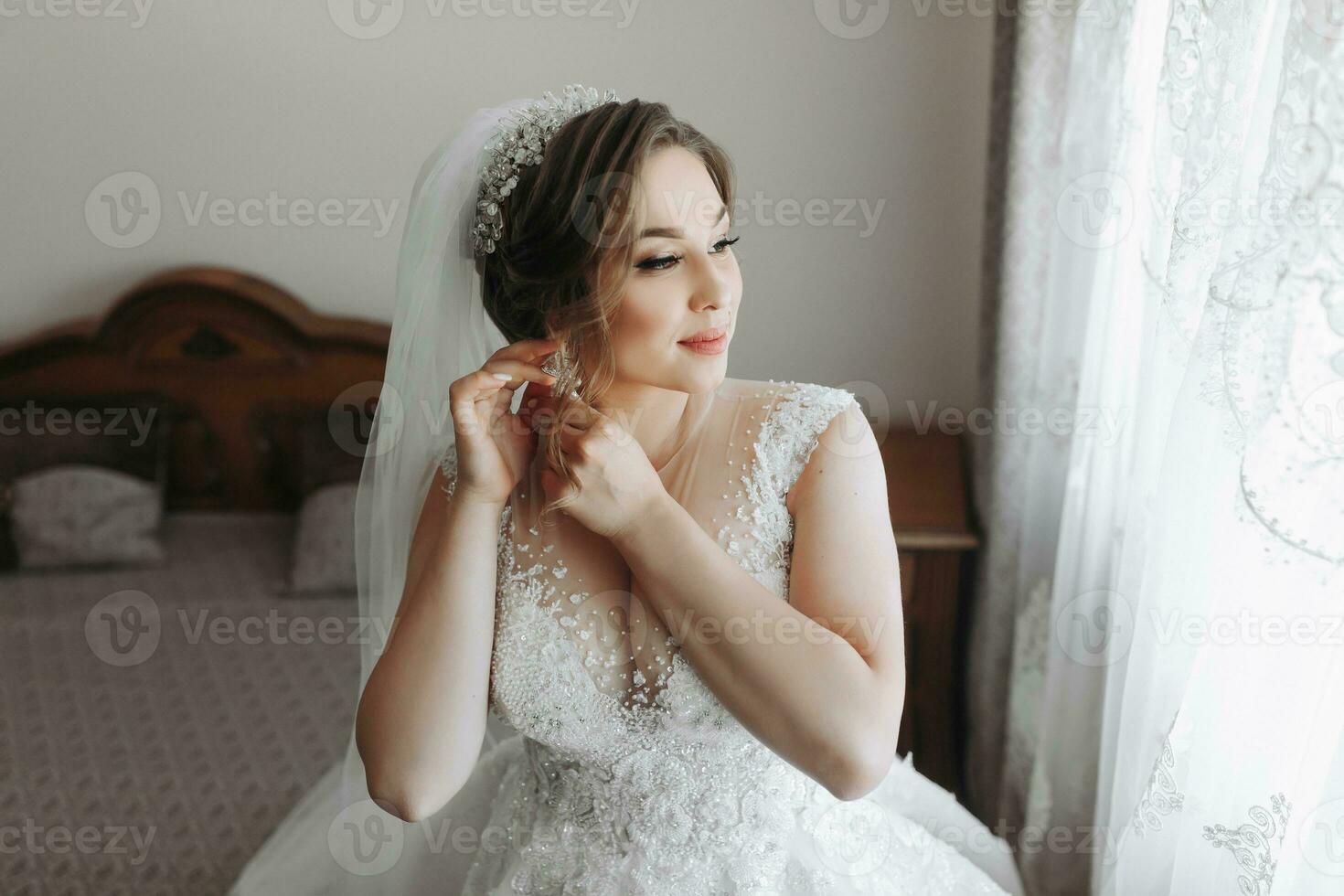 retrato de el novia. el novia es vestido en un elegante vestido, posando por el ventana vistiendo pendientes. tiara y joyas. abierto busto. Mañana de el novia foto