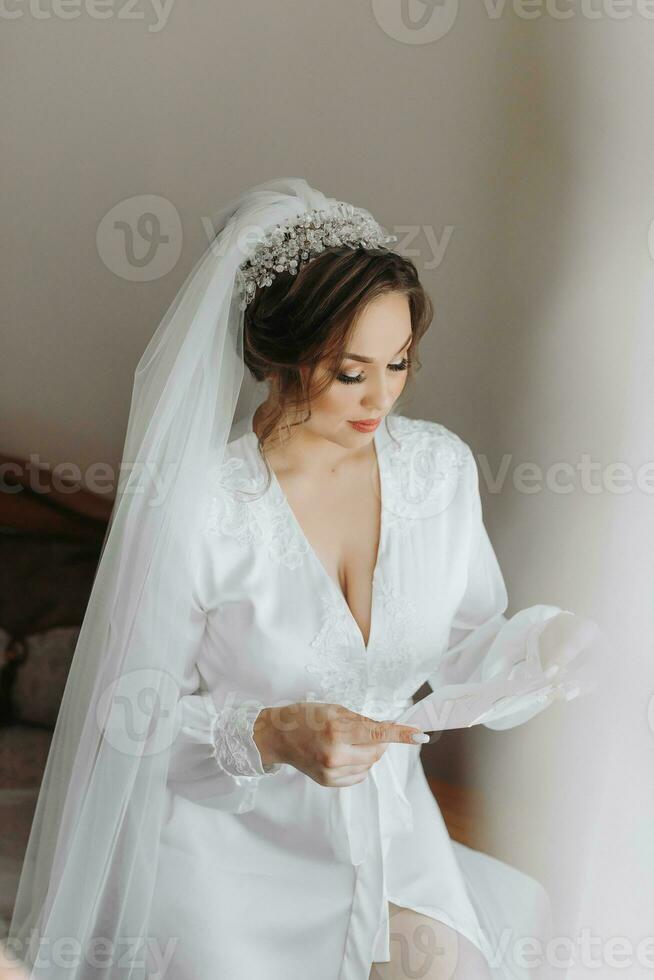 un hermosa novia en un túnica y lozano velo, con hermosa pelo y constituir, poses en su habitación. vendaje arriba y preparando para el Boda ceremonia foto