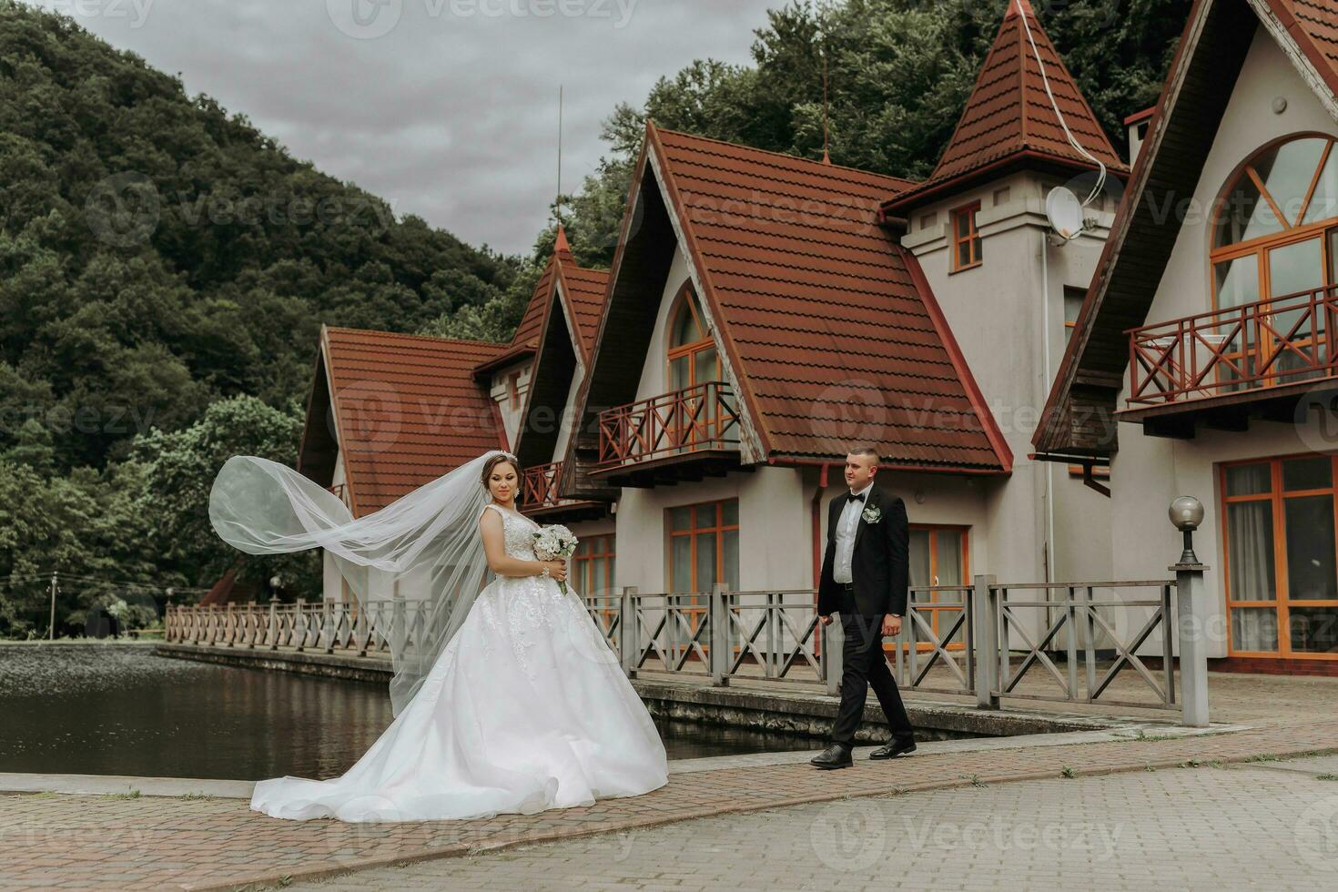 A brunette bride in a long dress and a groom in a classic suit are standing on a bridge near a lake against the background of a castle. A veil is thrown in the air photo
