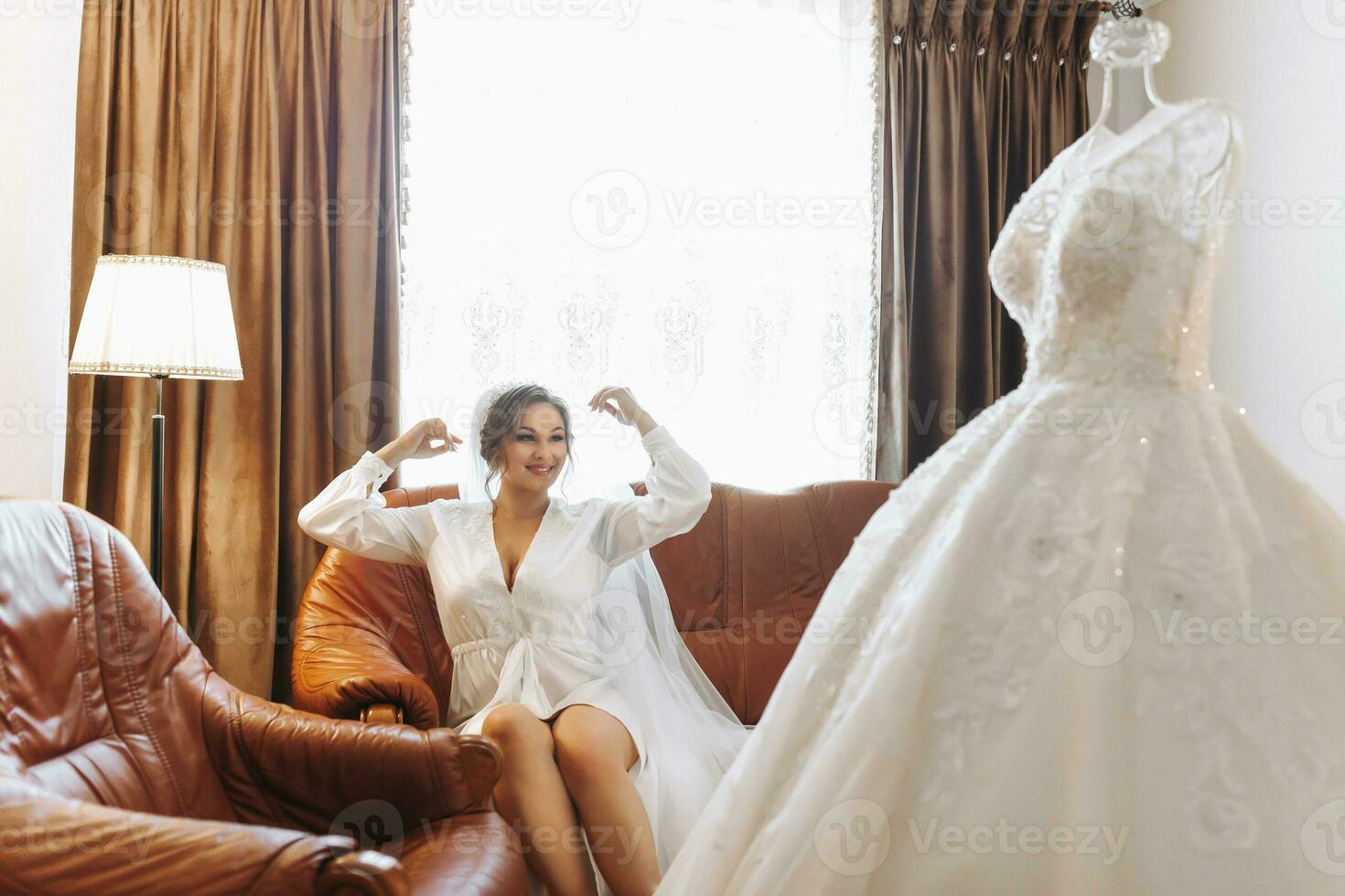 A beautiful bride in a robe with beautiful hair and makeup, sitting on a luxurious sofa, next to her dress on a mannequin. Dressing up and preparing for the wedding ceremony photo