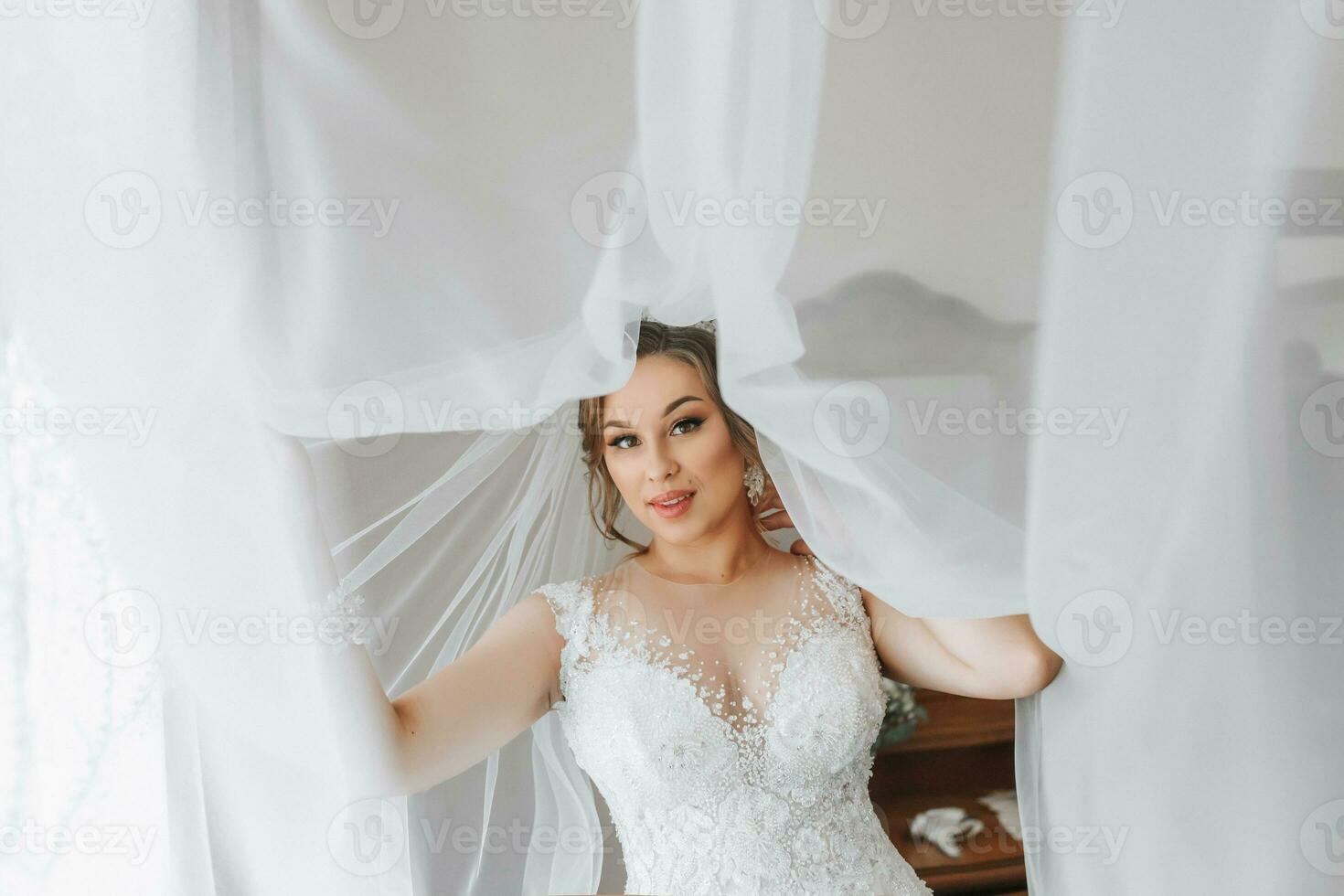 retrato de el novia. el novia en un velo, posando con un flor en el ojal, en un blanco antecedentes. hermosa pelo y constituir. abierto busto en el vestido. diadema con cristales foto