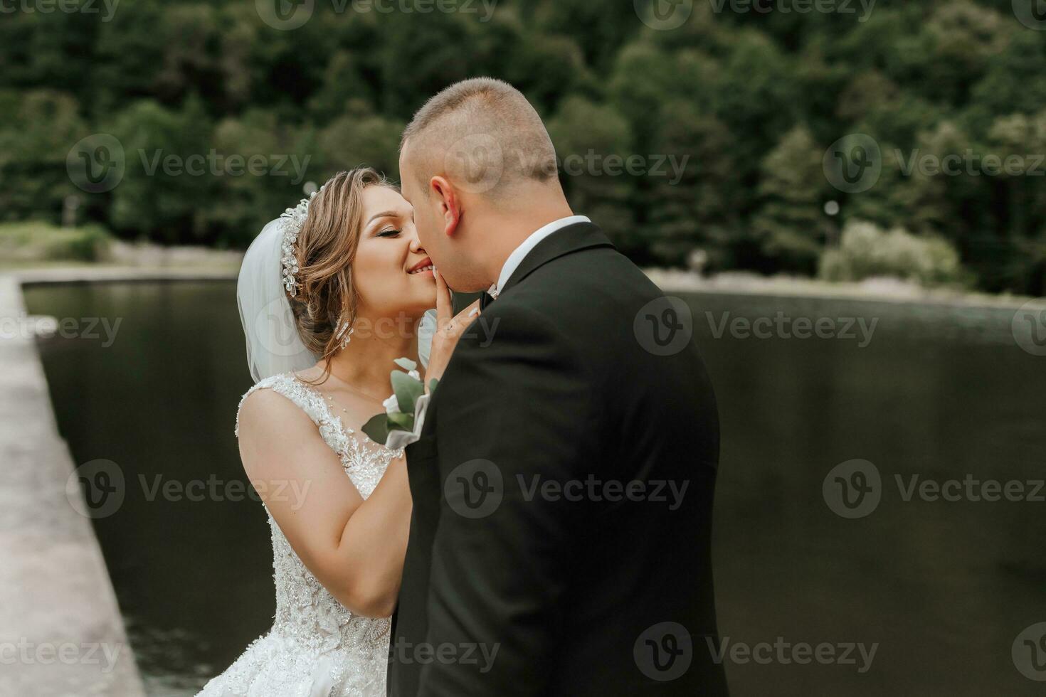A brunette bride in a long dress and a groom in a classic suit stand embracing on a bridge near a lake with a castle in the background. photo