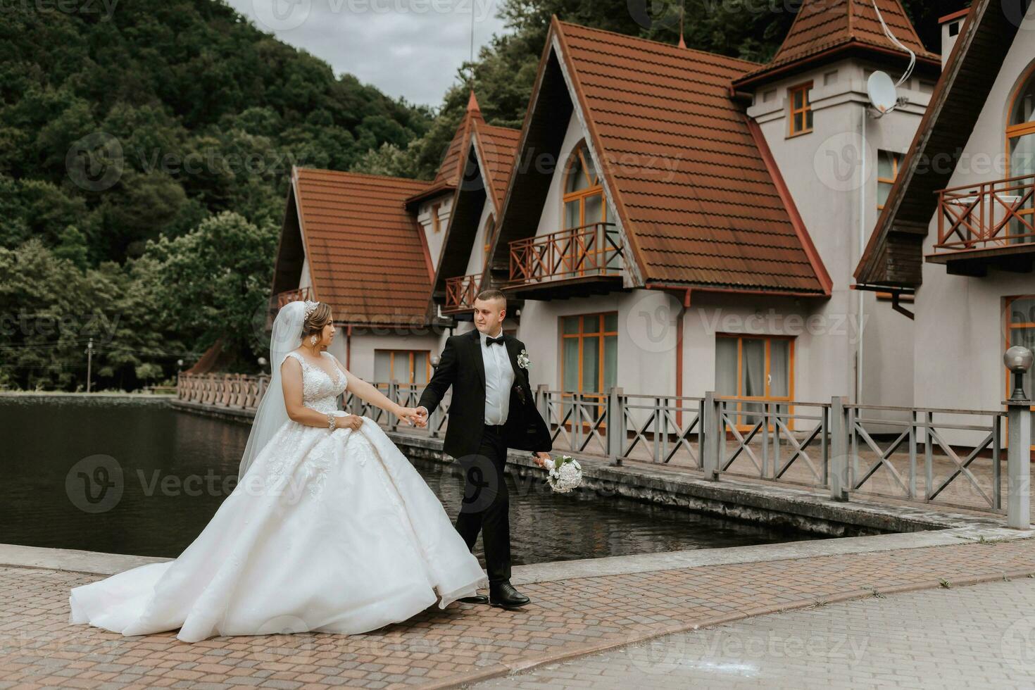 A brunette bride in a long dress and a groom in a classic suit are walking on a bridge near a lake against the background of a castle. a walk in nature. Wedding day photo