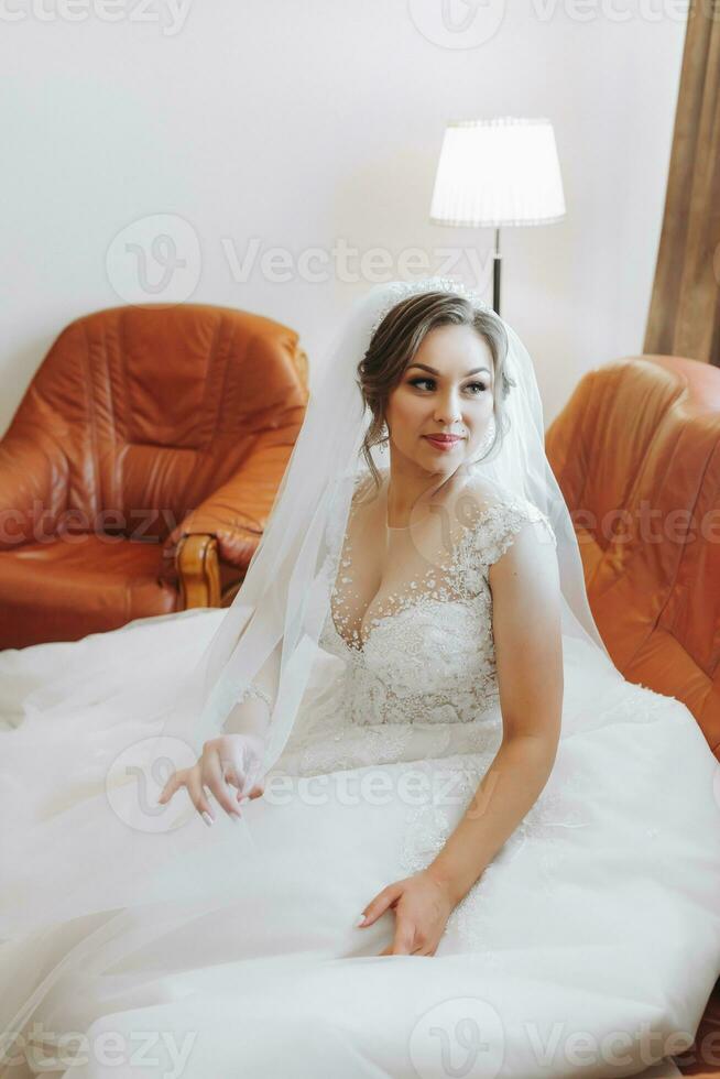 hermosa novia en un blanco vestir con hermosa pelo y maquillaje sentado en un lujoso sofá. vendaje arriba y preparando para el Boda ceremonia foto