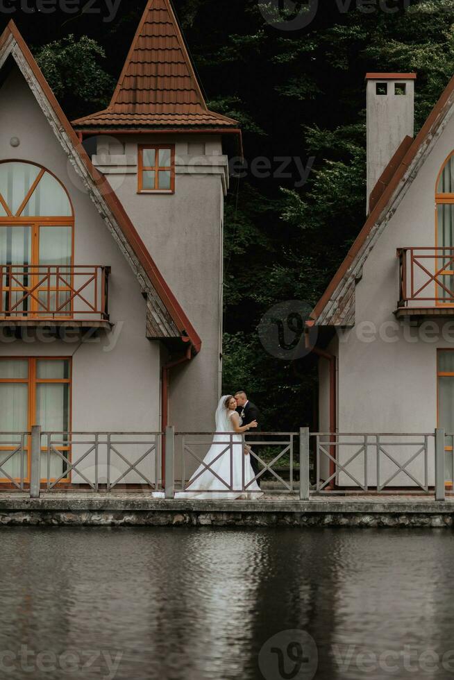 un morena novia en un largo vestir y un novio en un clásico traje son caminando en un puente cerca un lago en contra el antecedentes de un castillo. reflexión en el agua de el castillo. simétrico monitor foto