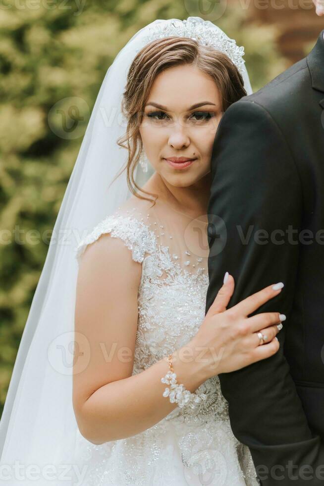 Fashionable groom and cute bride in white dress with crown hugging in park, garden, forest outdoors. Wedding photography, portrait of smiling newlyweds. photo