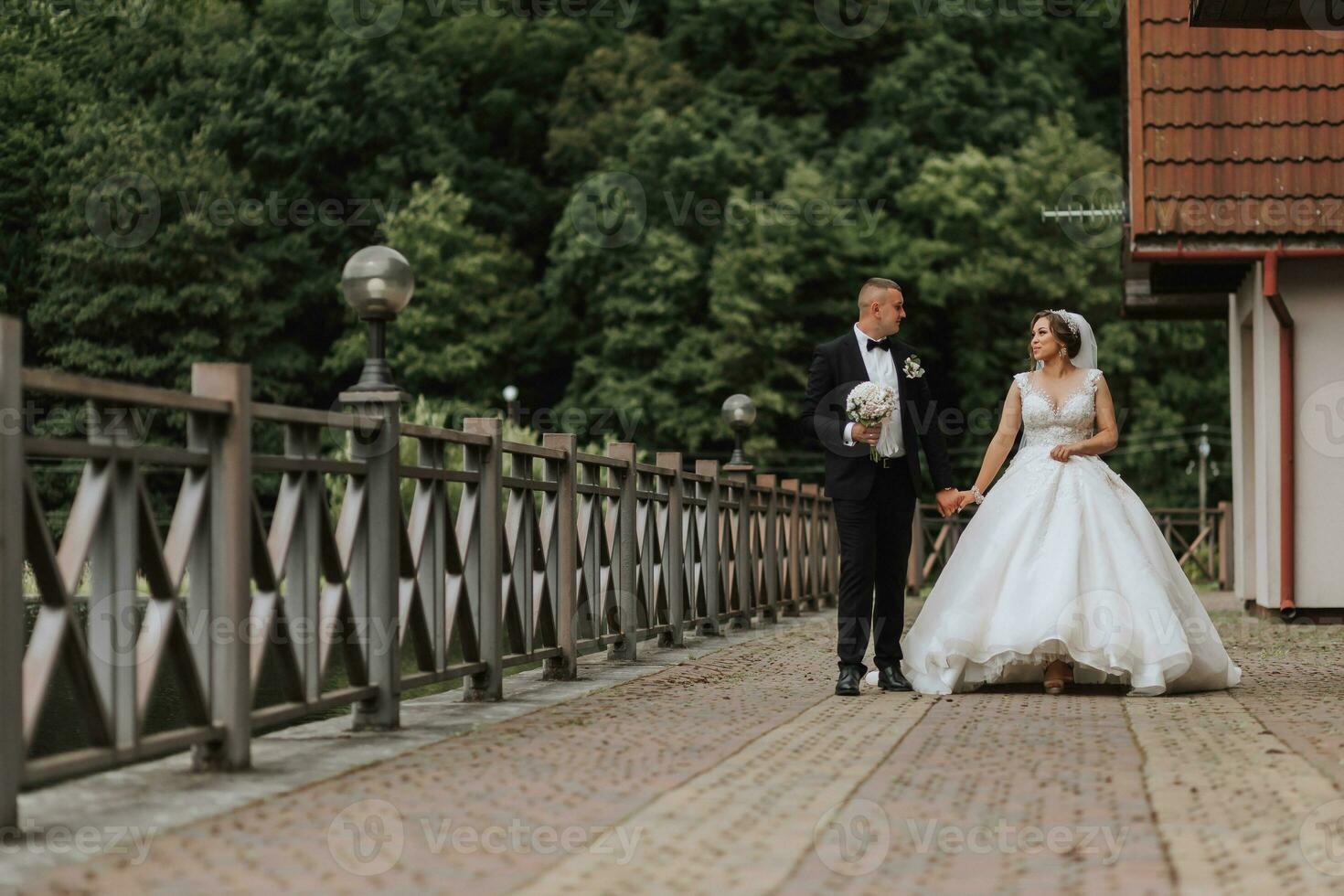 A brunette bride in a long dress and a groom in a classic suit are walking on a bridge near a lake against the background of a castle. a walk in nature. Wedding day photo