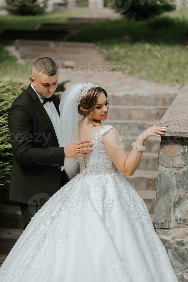 Wedding portrait. The bride in an elegant dress stands in front of the groom in a classic suit, against the background of green trees. Gentle touch. Summer wedding. A walk in nature photo