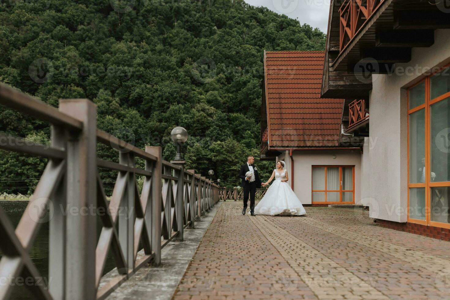 A brunette bride in a long dress and a groom in a classic suit are walking on a bridge near a lake against the background of a castle. a walk in nature. Wedding day photo