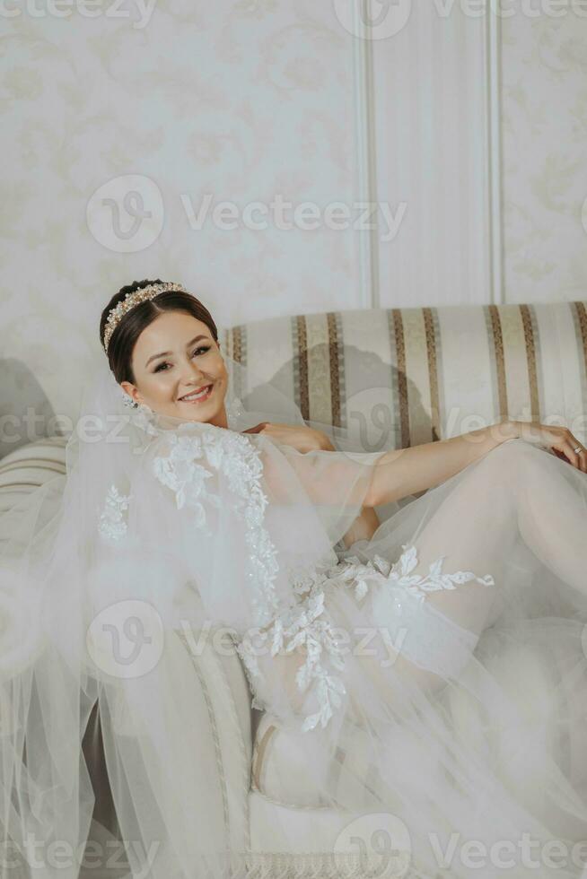 un hermosa niña con un Boda peinado y un tiara en su cabeza en un transparente túnica es preparando para un Boda en un hotel con un real interior. largo velo, natural luz, retrato foto