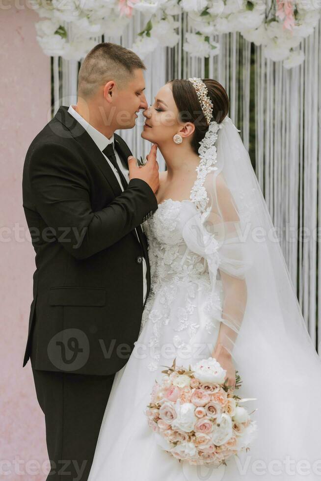 Attractive bride and groom at the ceremony on their wedding day with an arch made of pink and white flowers. Beautiful newlyweds, a young woman in a white dress with a long train, men in a black suit. photo