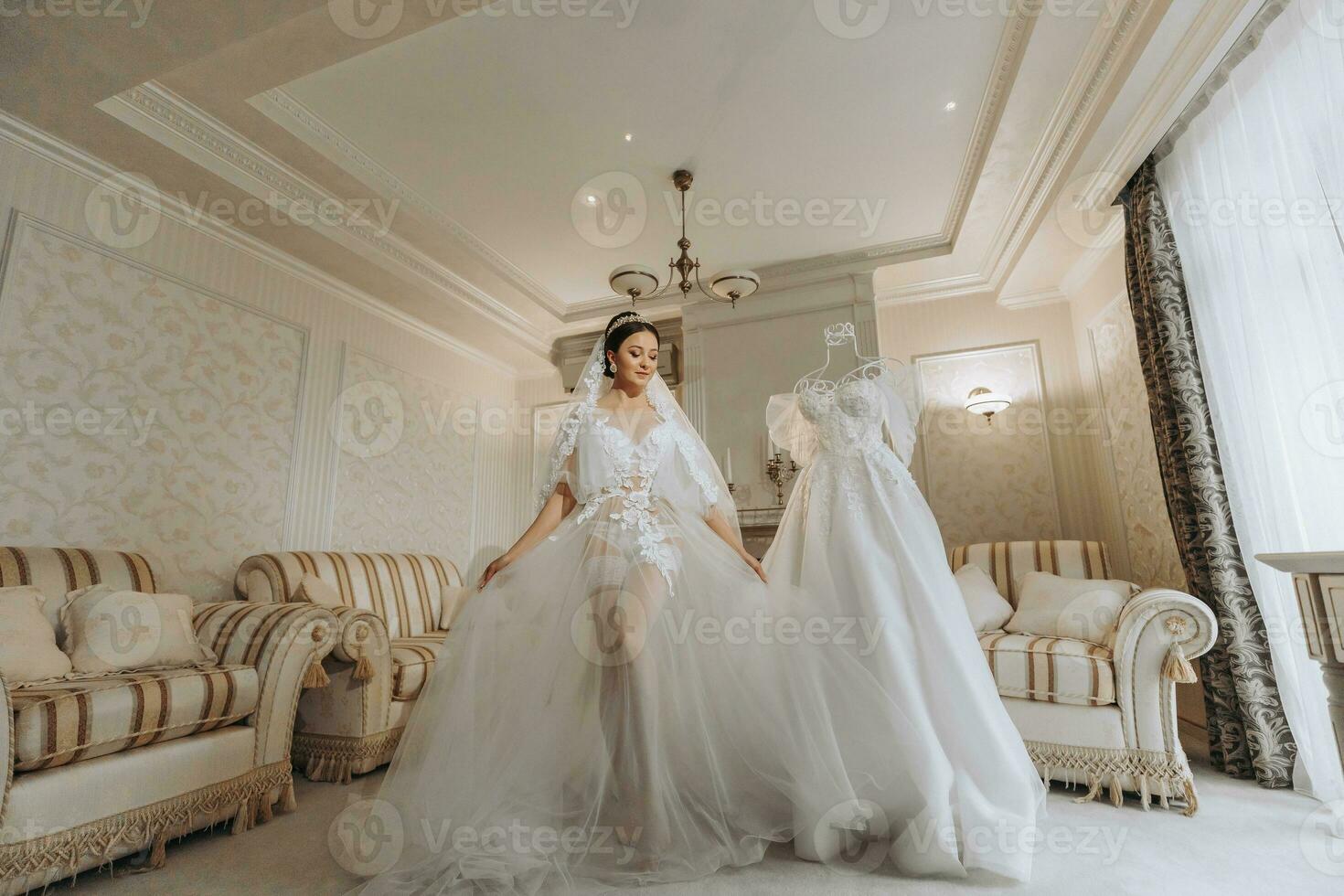 a beautiful girl with a wedding hairstyle in a transparent robe is preparing for a wedding in a hotel with a royal interior. The bride poses next to her wedding dress on a mannequin photo