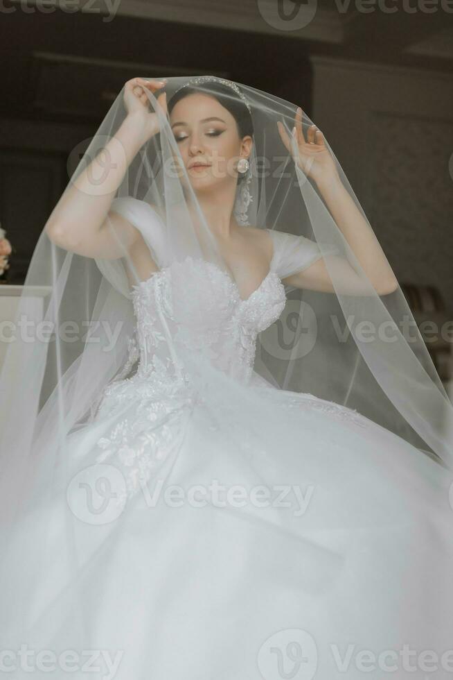 Preparation for the wedding. Beautiful young bride in white luxurious wedding dress, tiara on head, long veil in royal hotel room. Luxury smiling model photo