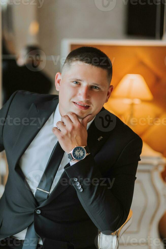 A stylish groom with a watch on his hand and a tie is sitting on a chair in an expensive hotel room. Groom's morning. The groom is getting ready in the morning before the wedding ceremony. photo