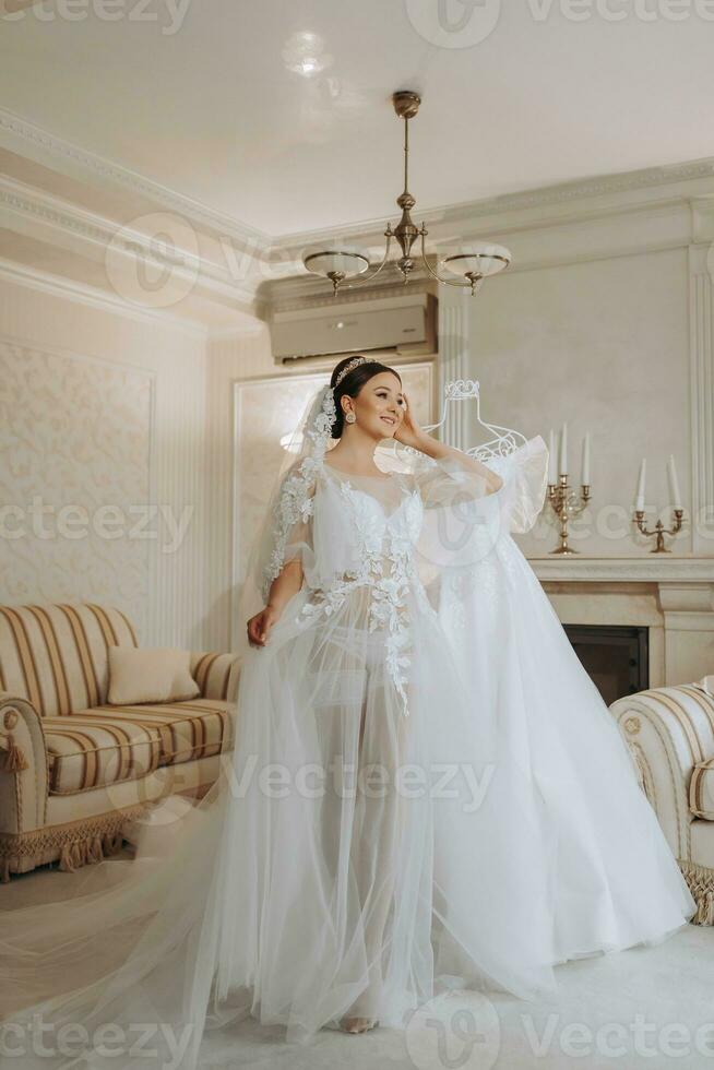 a beautiful girl with a wedding hairstyle in a transparent robe is preparing for a wedding in a hotel with a royal interior. The bride poses next to her wedding dress on a mannequin photo