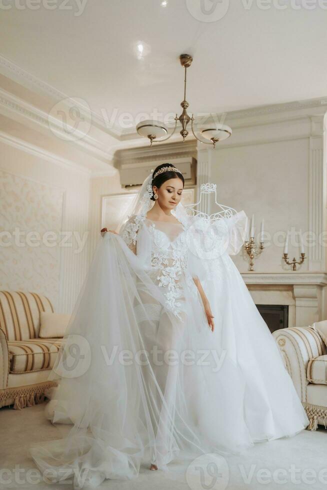 a beautiful girl with a wedding hairstyle in a transparent robe is preparing for a wedding in a hotel with a royal interior. The bride poses next to her wedding dress on a mannequin photo