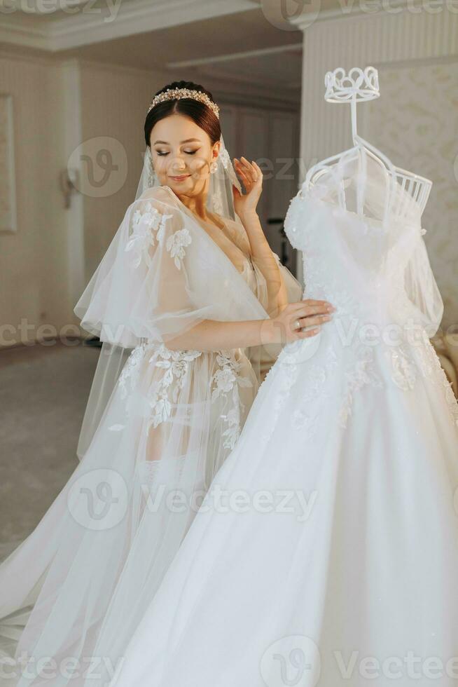 a beautiful girl with a wedding hairstyle in a transparent robe is preparing for a wedding in a hotel with a royal interior. The bride poses next to her wedding dress on a mannequin photo