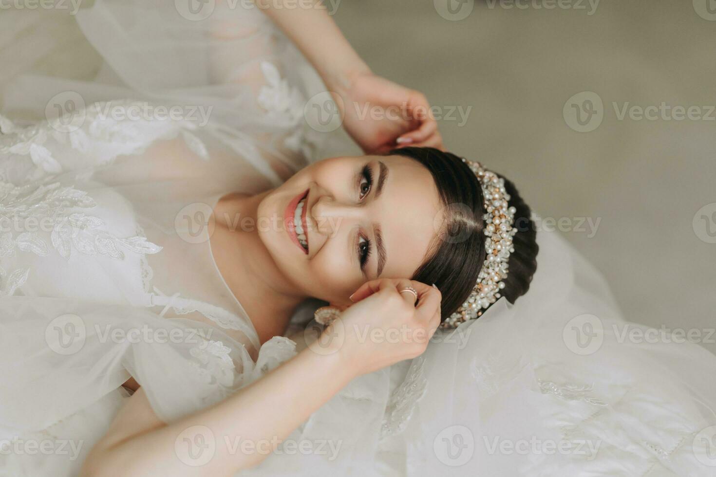 a beautiful girl with a wedding hairstyle and a tiara on her head in a transparent robe, lying on a bed in a hotel with a royal interior. Long veil, natural light, portrait photo