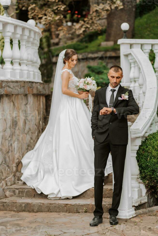 Portrait of a young bride in a white dress and a brunette groom in a suit on the stairs near the old city park. The man in the foreground. A beautiful and romantic wedding, a happy couple photo