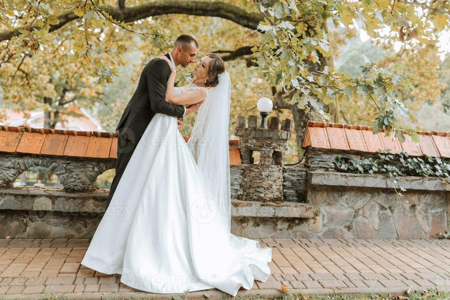 Fashionable groom and cute brunette bride in white dress with professional make-up and bouquet of flowers are hugging, laughing in park, garden, forest outdoors. Wedding photography photo