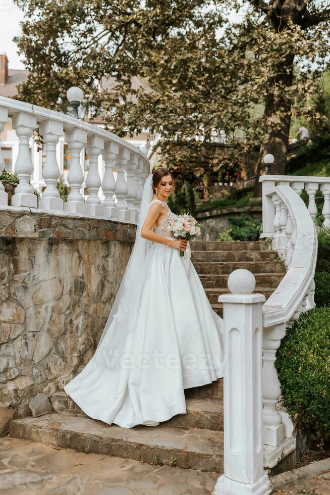 A beautiful brunette bride in a white long dress with a bouquet of flowers stands on the stairs against the background of tall trees, outdoors in the park. photo