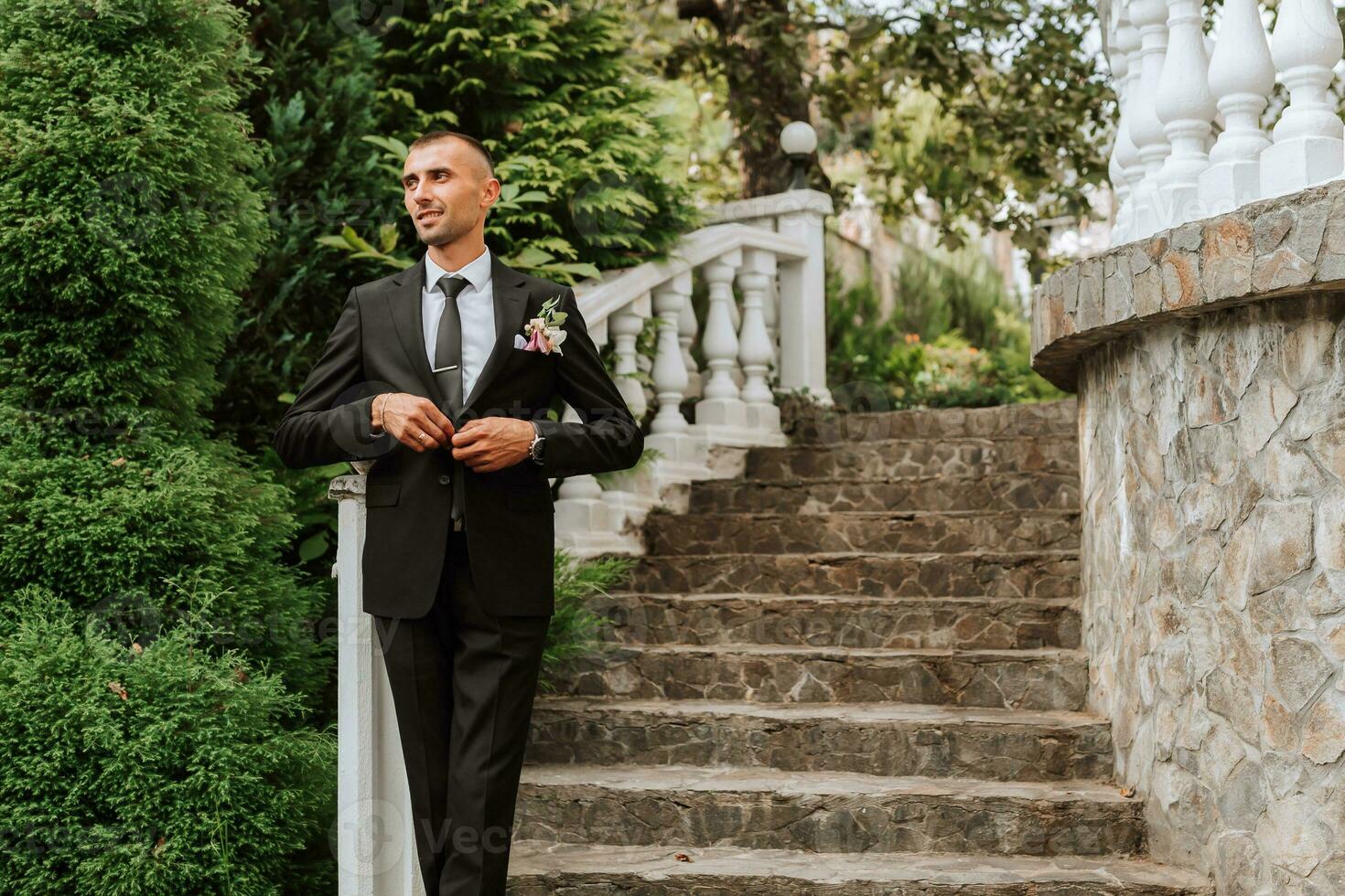 A man in a classic black suit and white shirt stands and poses near the stone steps. A stylish watch. Men's style. Fashion. Business. photo