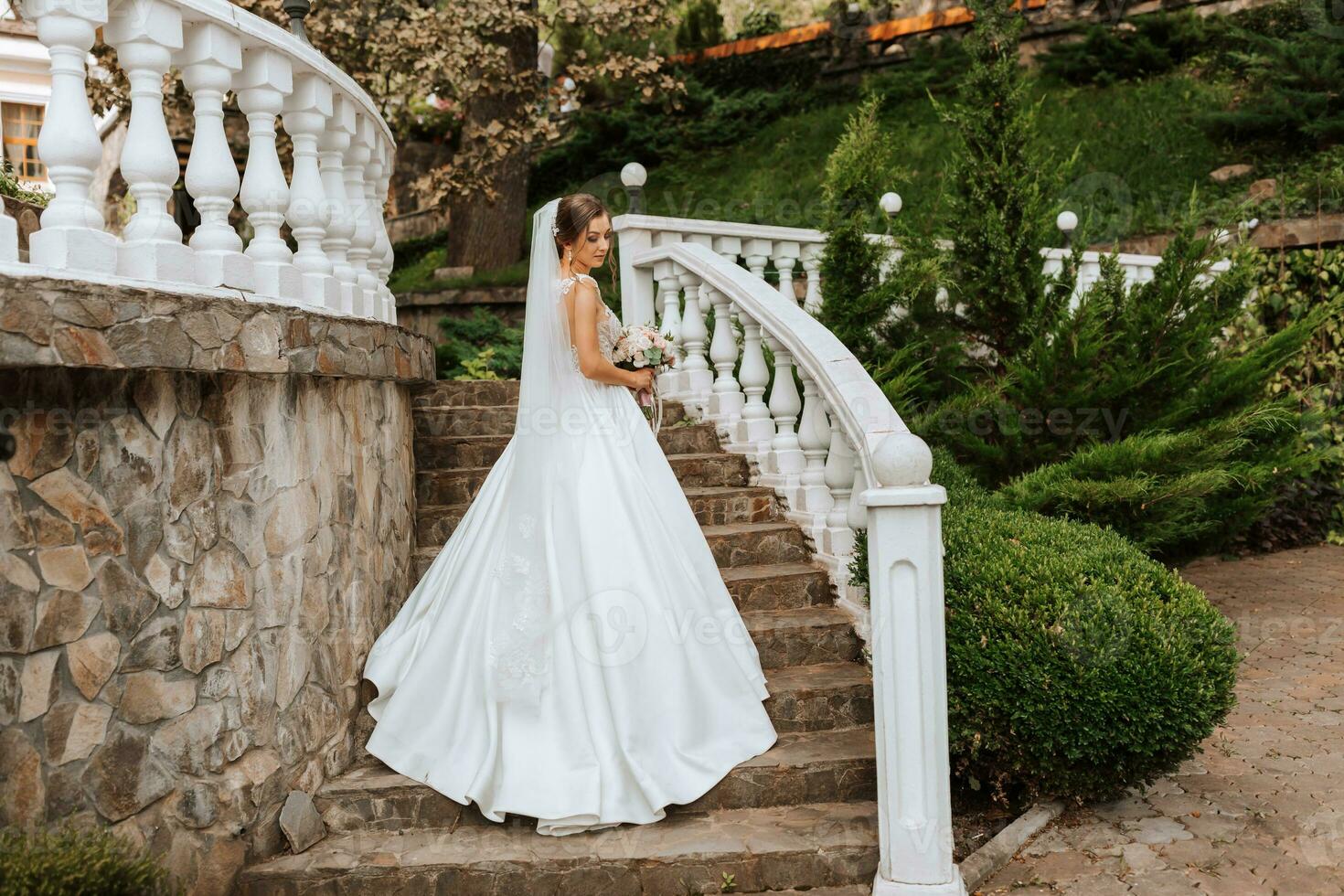 A beautiful brunette bride in a white long dress with a bouquet of flowers stands on the stairs against the background of tall trees, outdoors in the park. photo