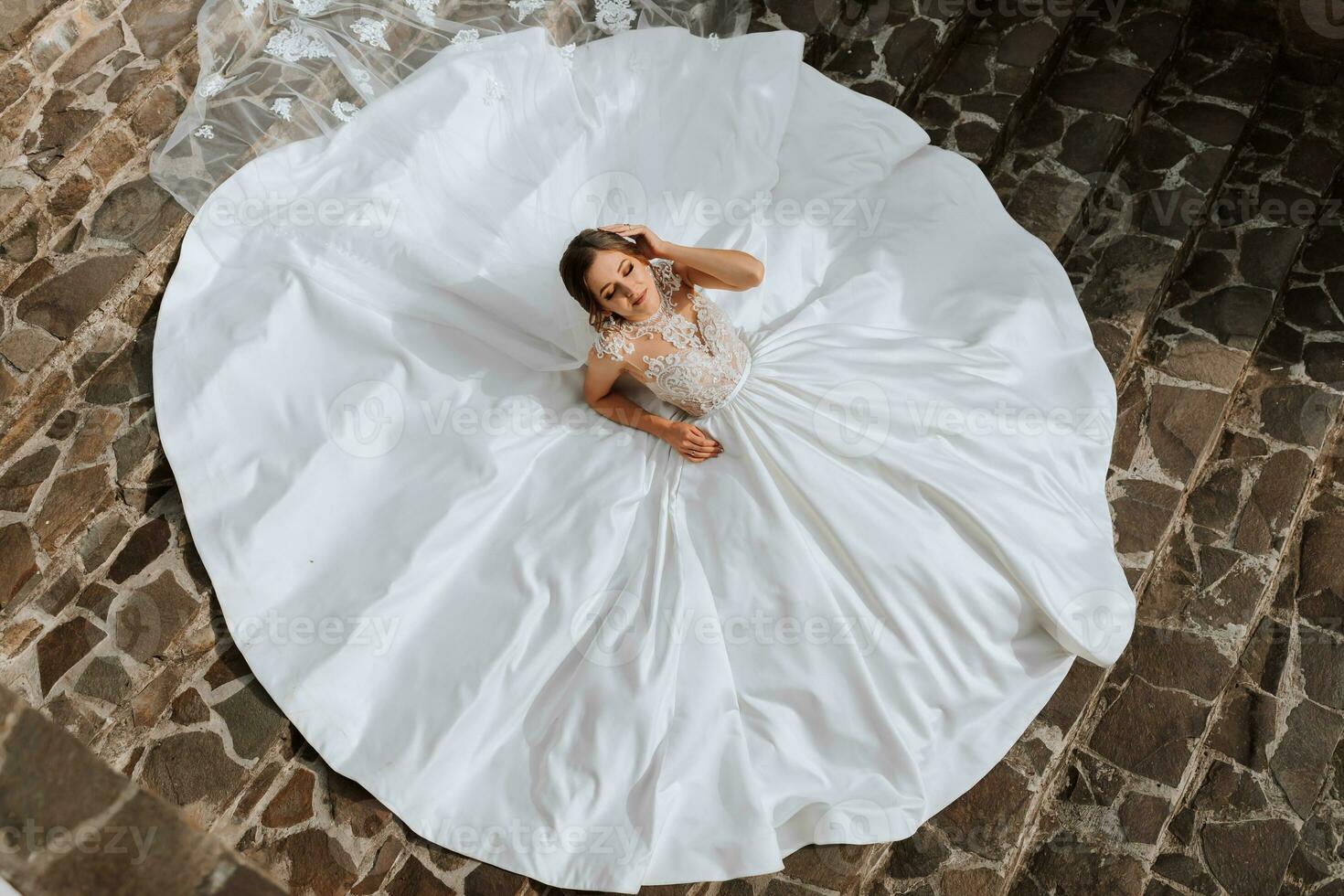 young beautiful bride in a wedding dress with open shoulders and a crown on her head is lying on stone stairs, fashion shot under harsh sunlight. The photo was taken from above