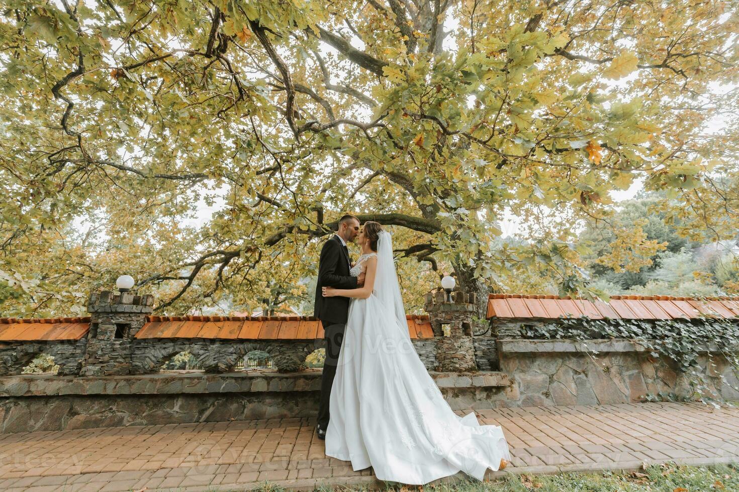 Fashionable groom and cute brunette bride in white dress with professional make-up and bouquet of flowers are hugging, laughing in park, garden, forest outdoors. Wedding photography photo