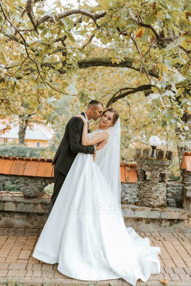 Fashionable groom and cute brunette bride in white dress with professional make-up and bouquet of flowers are hugging, laughing in park, garden, forest outdoors. Wedding photography photo